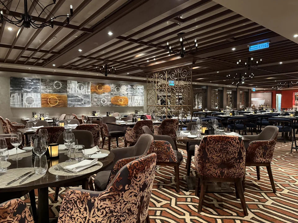 Indretningen i La Cucina på Norwegian Escape med træbjælkeloft, smedejernslysekroner og dækkede borde