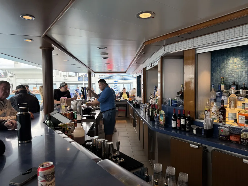 Udendørs bar ved pooldækket på Norwegian Escape med bartender og gæster på havdagen