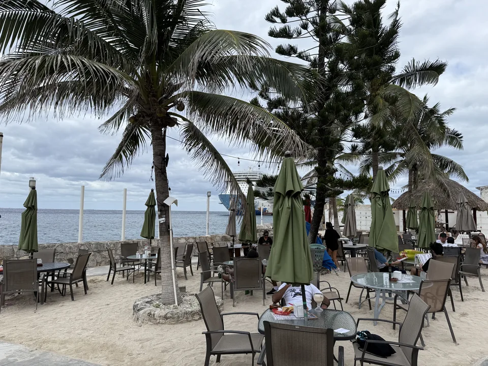 Strandbar med palmer og parasoller på sandet ved vandet på Cozumel med Norwegian Escape i baggrunden