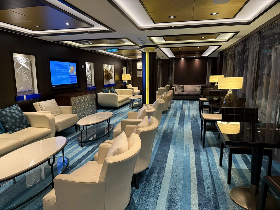 Rolig lounge med lyse læderlænestole, lamper og blåt stribet tæppe om bord på Norwegian Escape