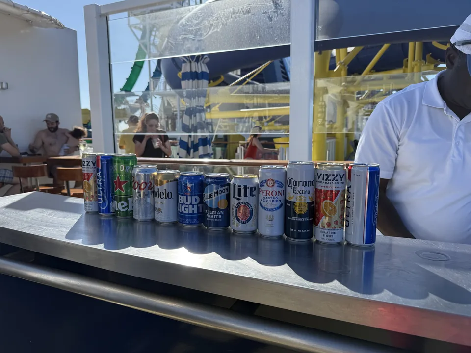Række af øldåser på baren ved pooldækket på Norwegian Escape med vandrutsjebaner i baggrunden