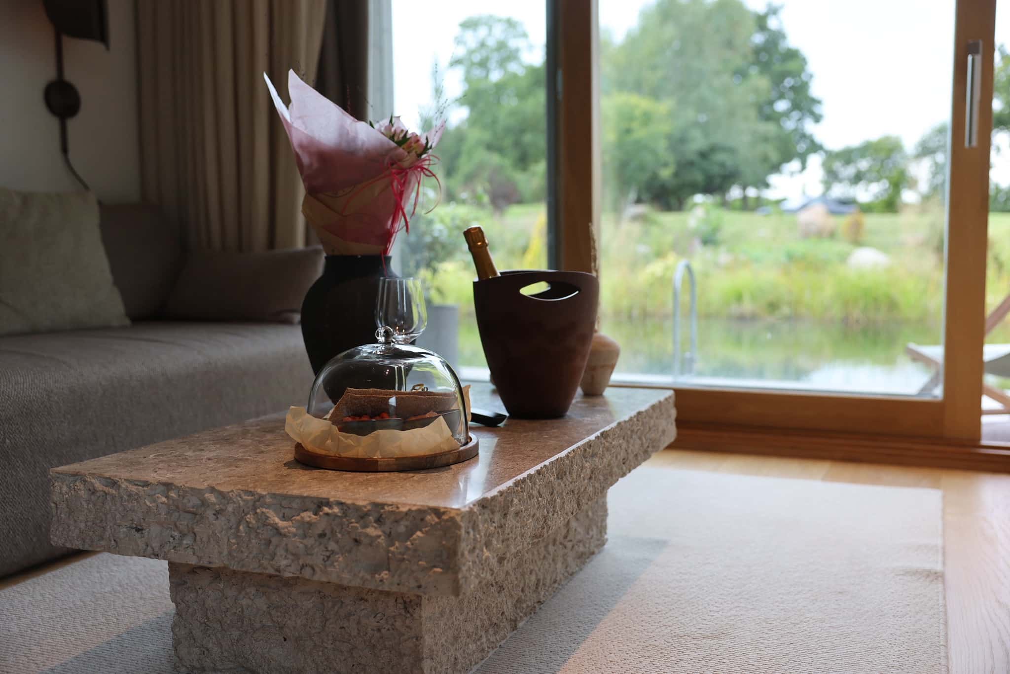 Stenbord med champagnekøler, glasklokke med chokolade og blomster foran vinduer mod sø