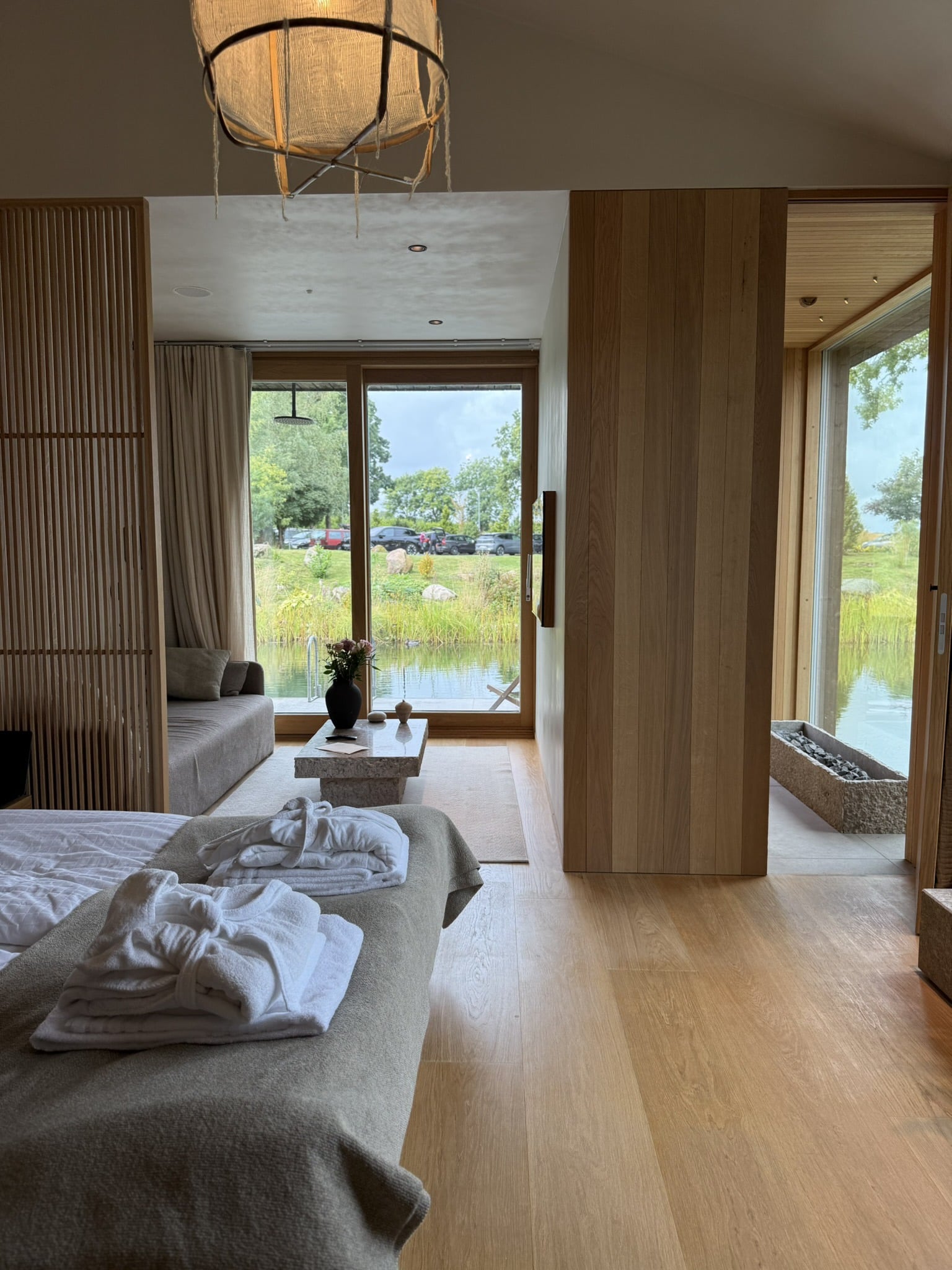 Hotelværelse i lyst træ med seng, sofaområde og glaspartier mod sø