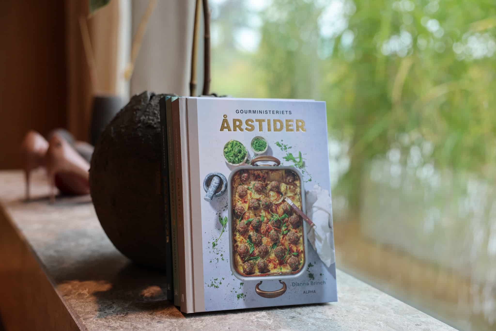 Kogebogen Gourministeriets Årstider står på vindueskarm med sløret grøn baggrund