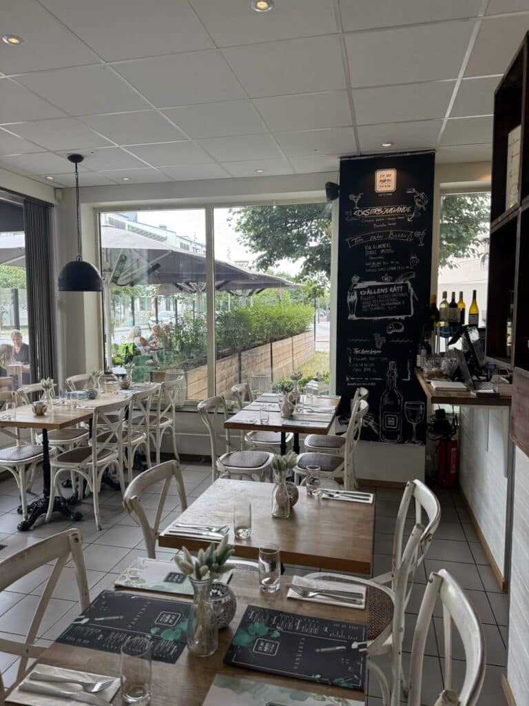 Restaurantlokale med hvide stole, træborde, tavle med tilbud og store vinduer.