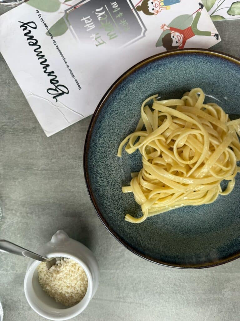 Blå skål med tagliatelle uden sauce og en lille kande revet parmesan.
