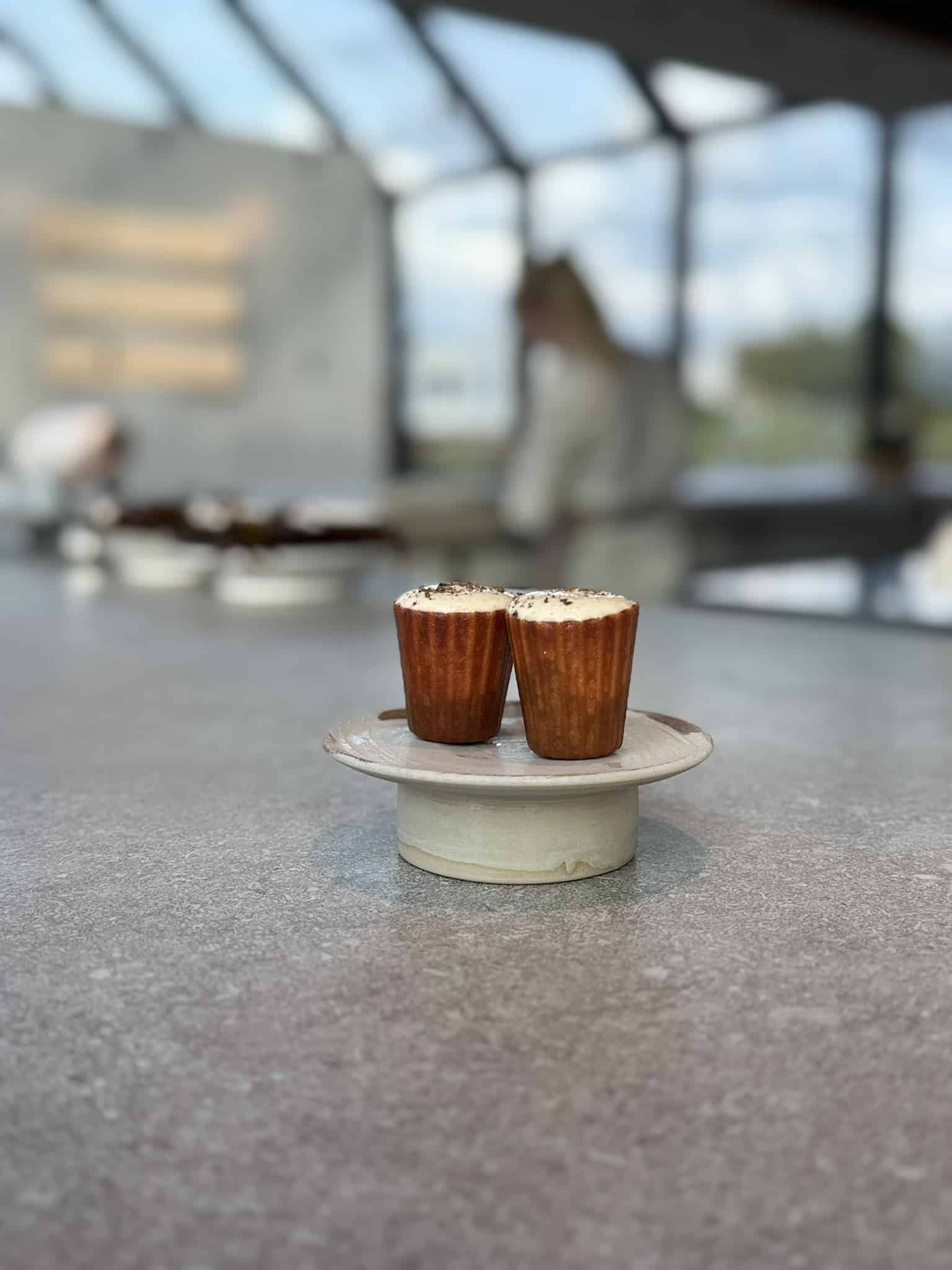 To små brune canelé-formede kager på keramikfad foran åbent køkken.