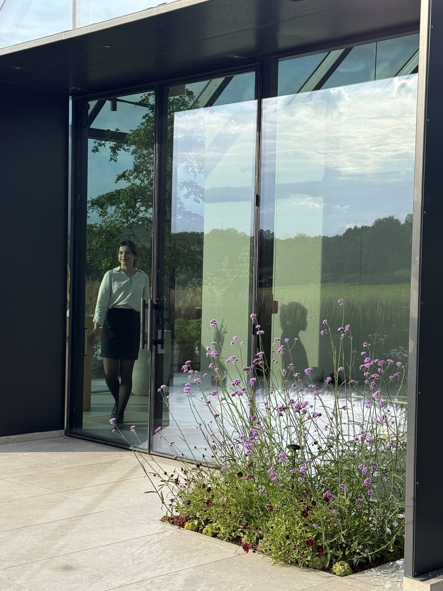 Glasfacade med indgangsdør, kvinde i hvid skjorte indenfor, lilla blomster foran.