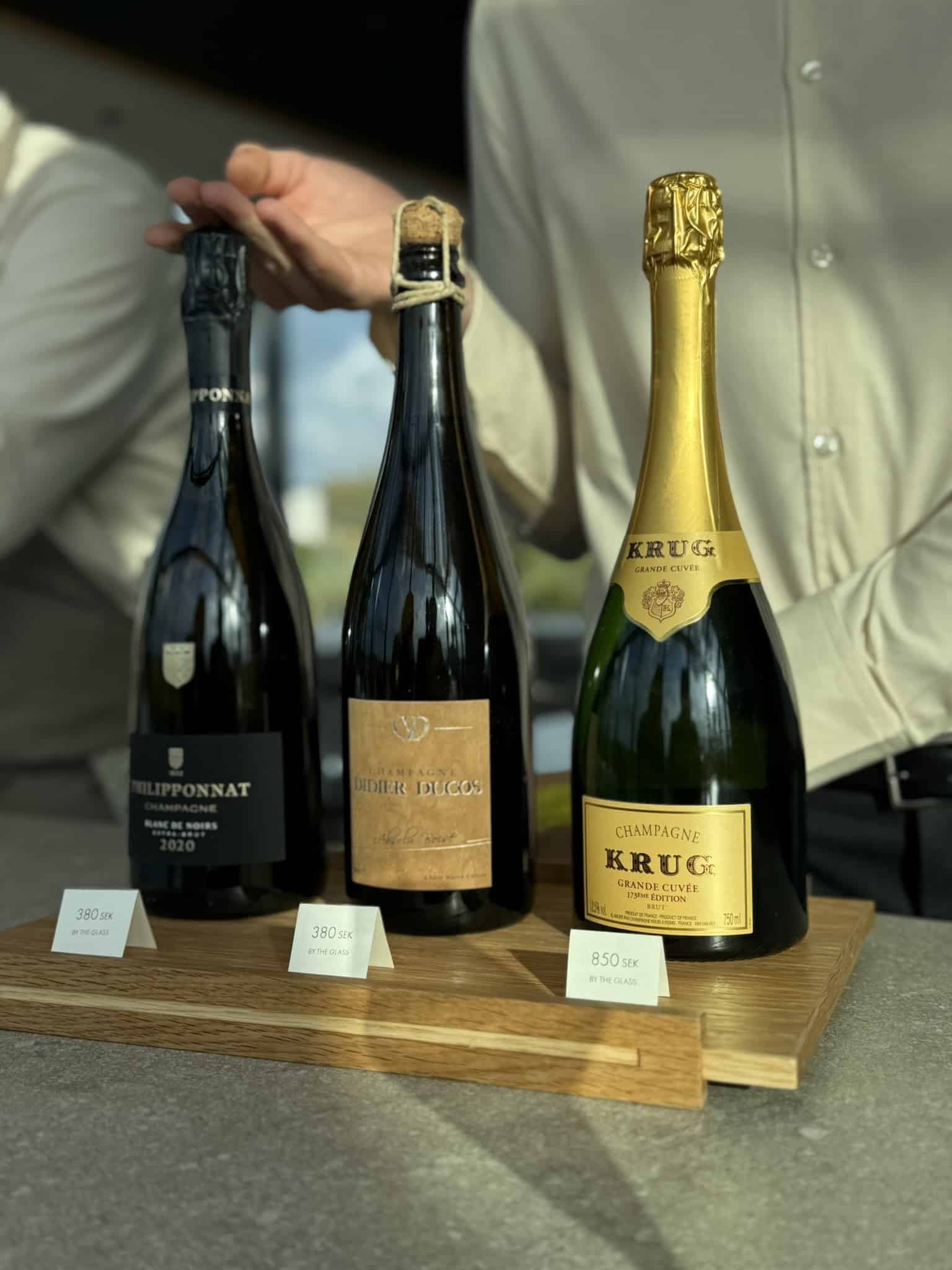 Tre champagneflasker på træplade med prisskilte: Philipponnat, Didier Ducos, Krug.
