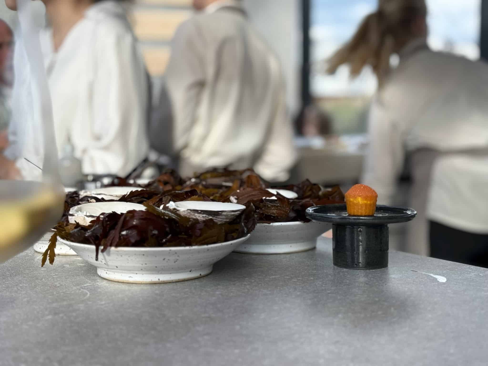 Skåle med tang og små hvide skaller samt lille orange kage på sort stand.