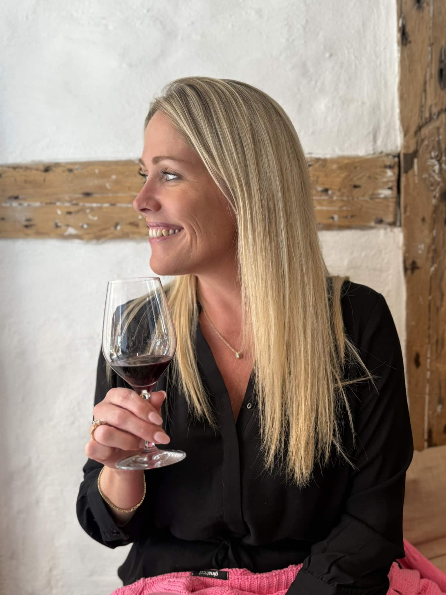 Smilende kvinde i sort skjorte holder et glas rødvin foran bindingsværksvæg