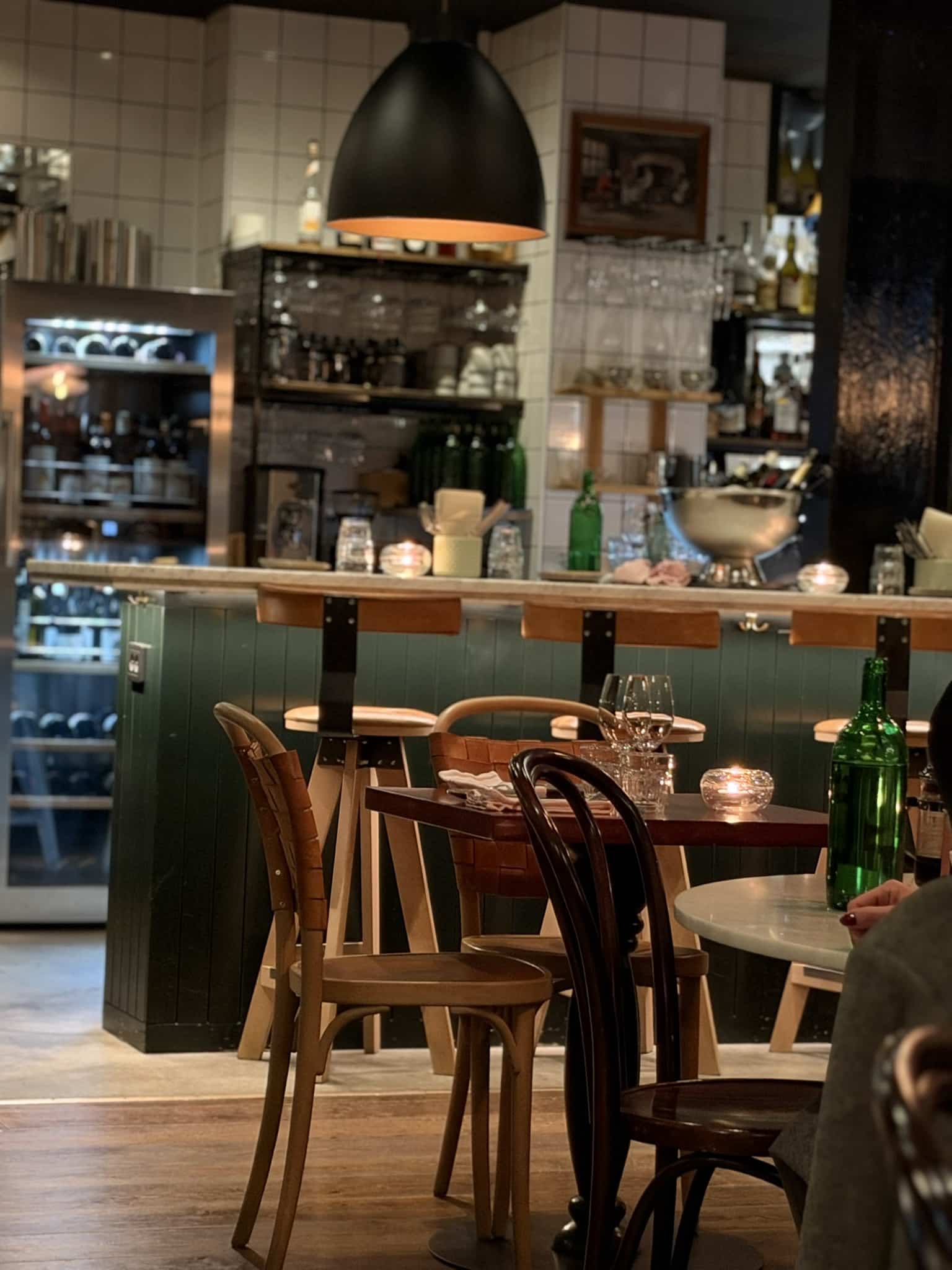Restaurantlokale med bardisk, grøn træbeklædning, hvide fliser, vinkøleskab og træstole