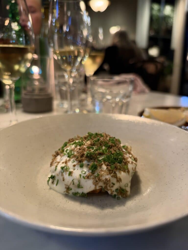 Tærte med creme, trøffel og purløg foran vinglas og sløret restaurantlokale