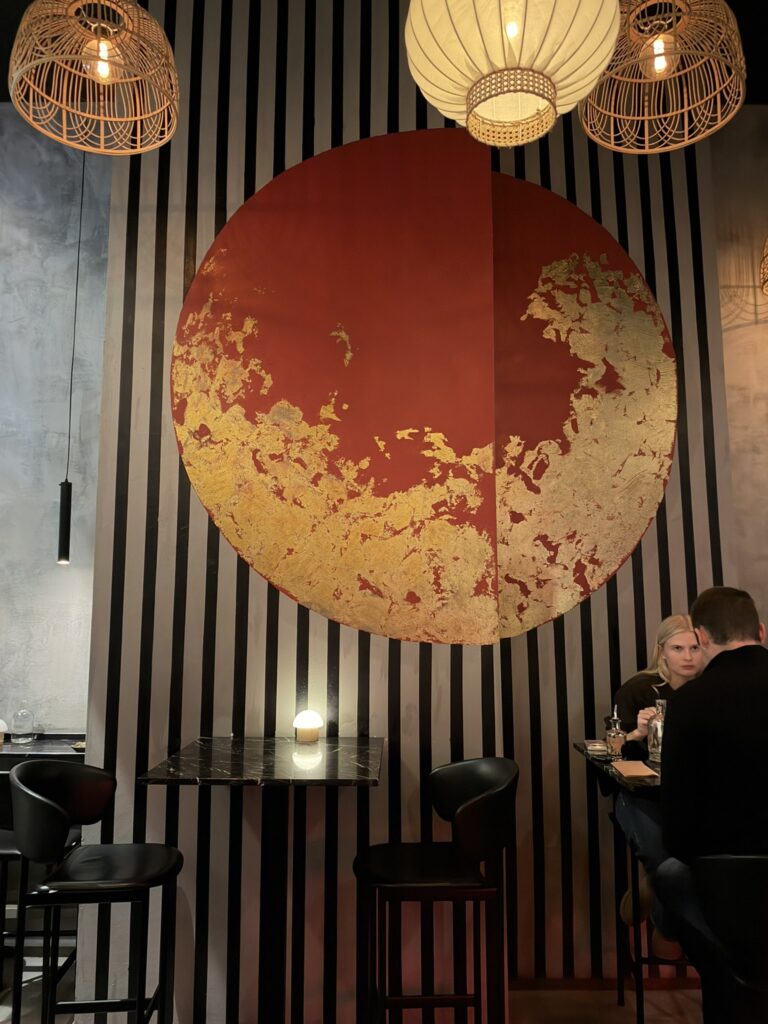 Restaurantlokale med rød cirkel på stribet væg, barstole og rattanlamper.