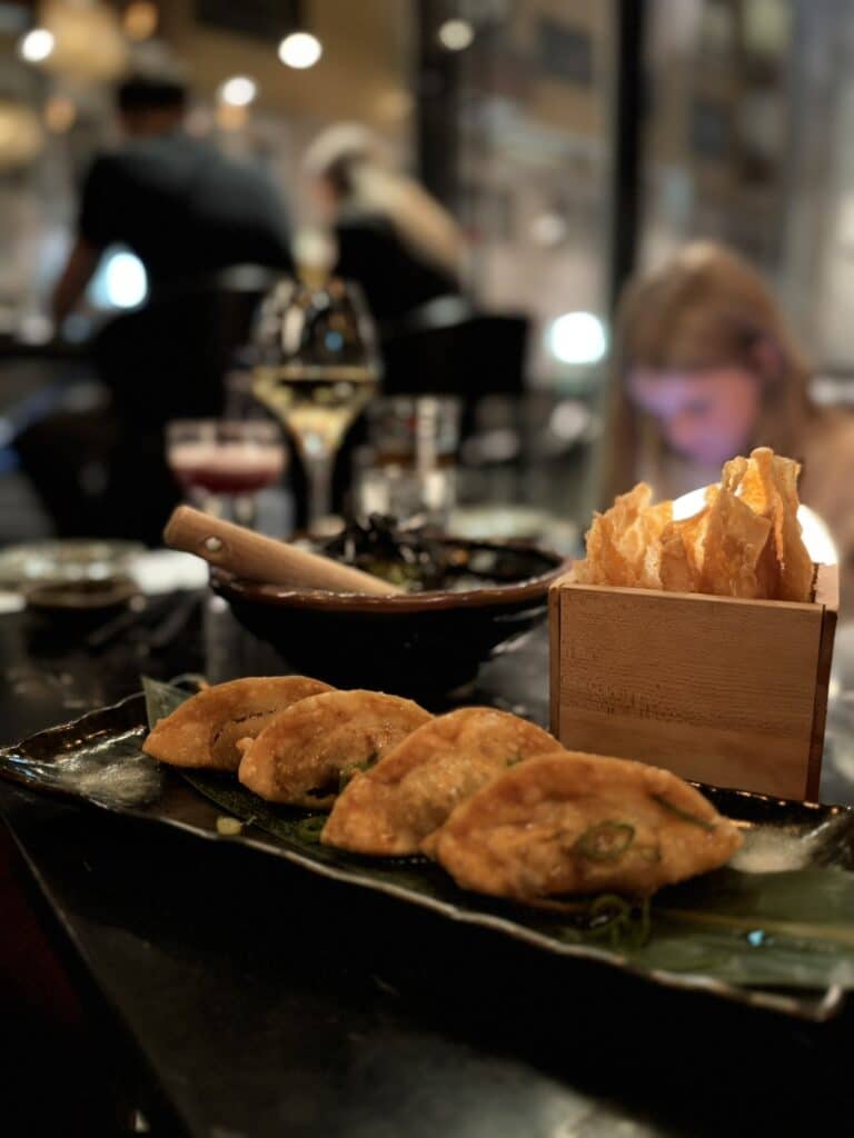 Fire stegte gyoza på aflangt fad med forårsløg, chips i baggrunden.