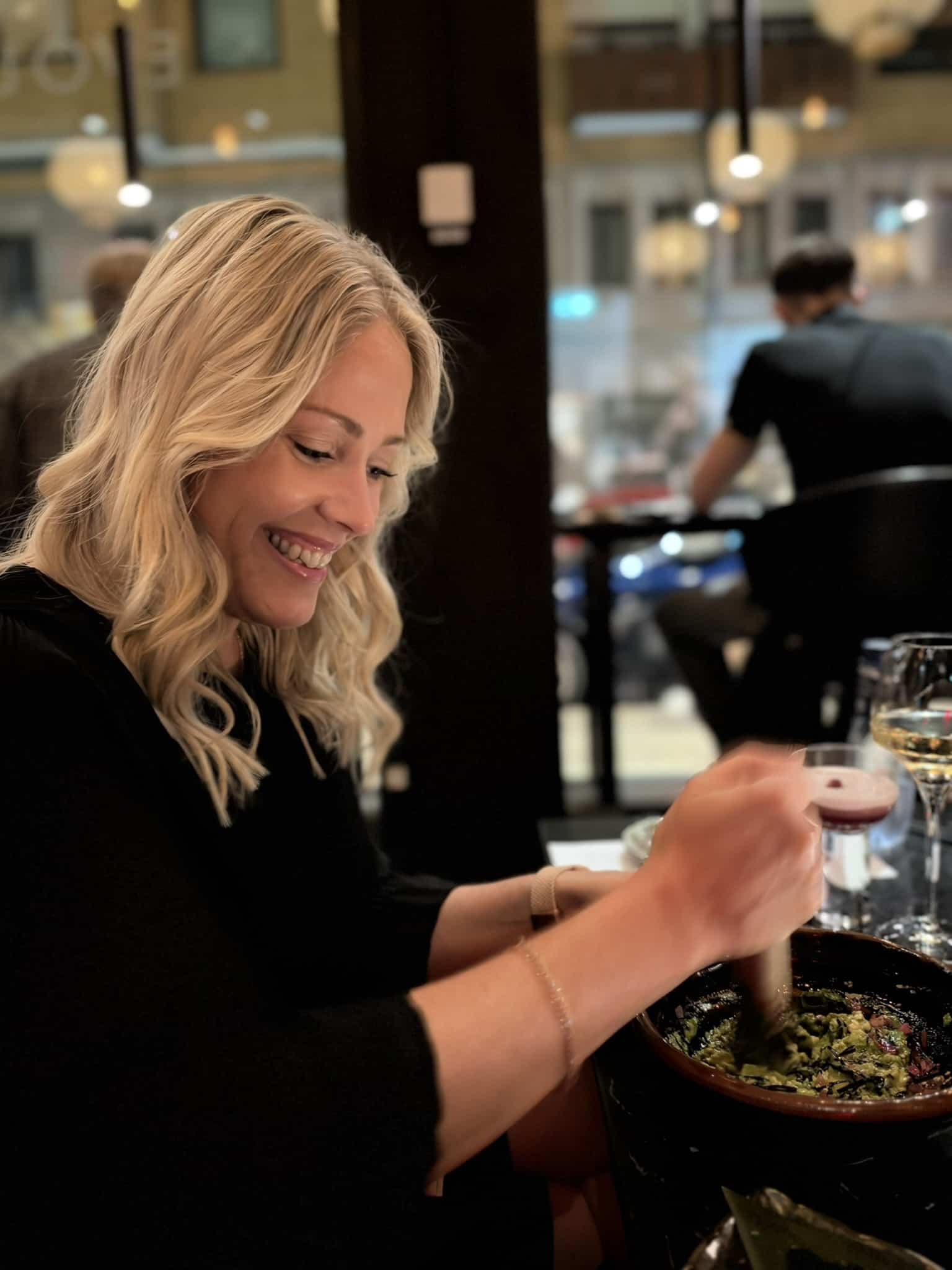 Smilende kvinde støder guacamole i morter ved bord med cocktail og hvidvin