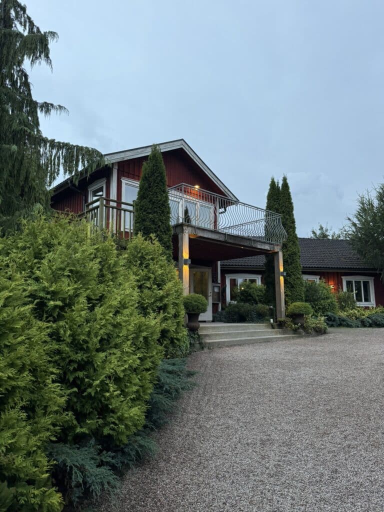 Rød træbygning med balkon, grusplads og klippede thujaer i skumring