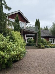 Rød træbygning med balkon, cypresser og grusbelagt gårdsplads foran.