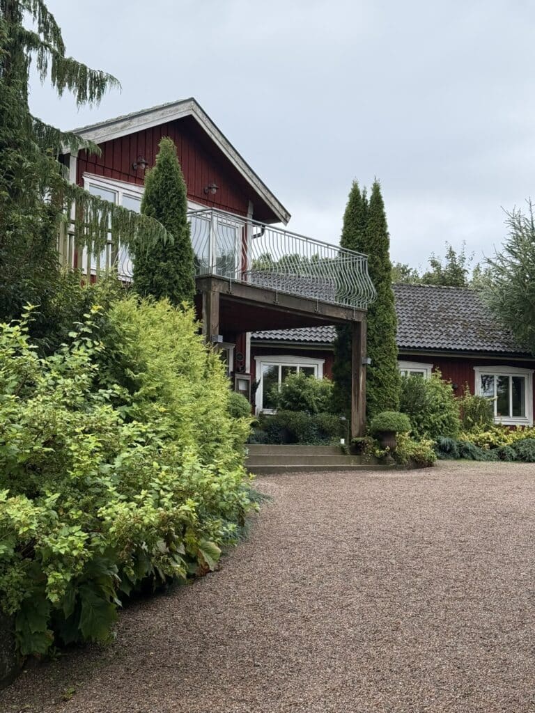 Rød træbygning med balkon, cypresser og grusbelagt gårdsplads foran.