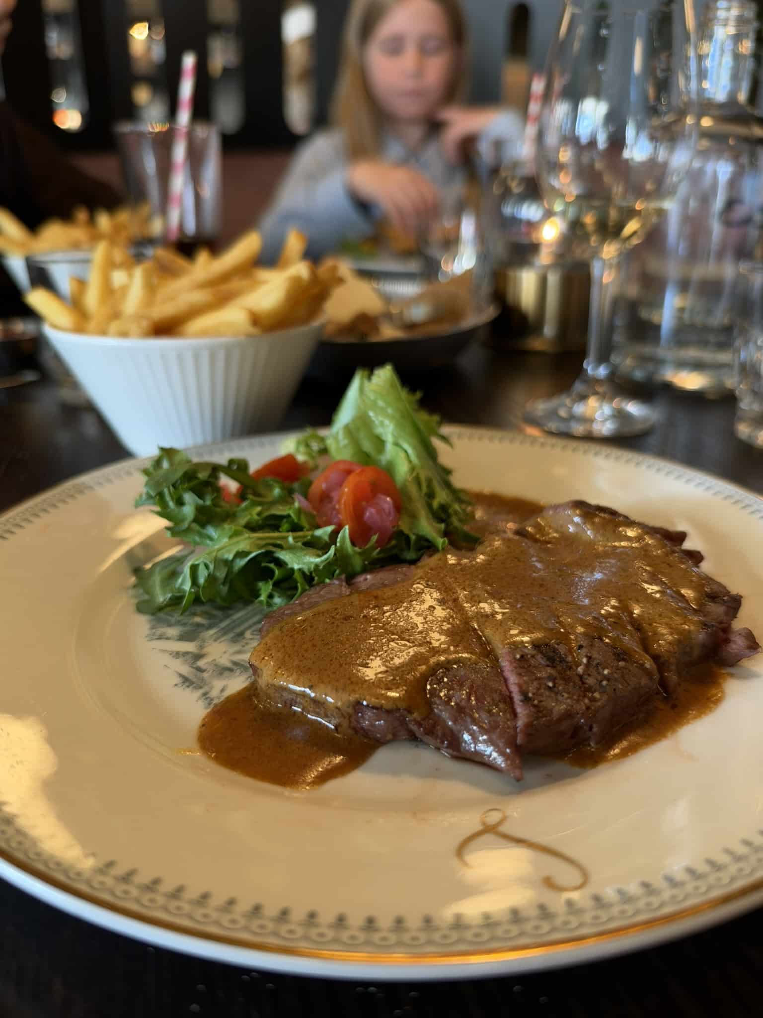 Steak med pebersauce og salat, pommes frites og barn i baggrunden
