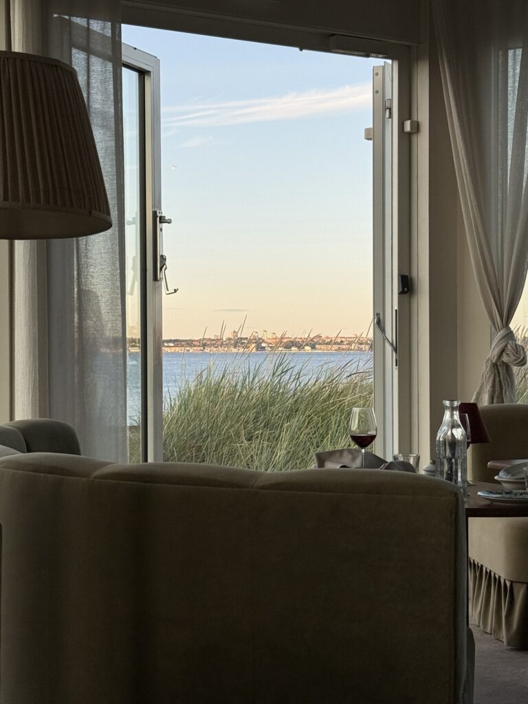 Åben terrassedør med udsigt over strandgræs og hav mod byens kyst