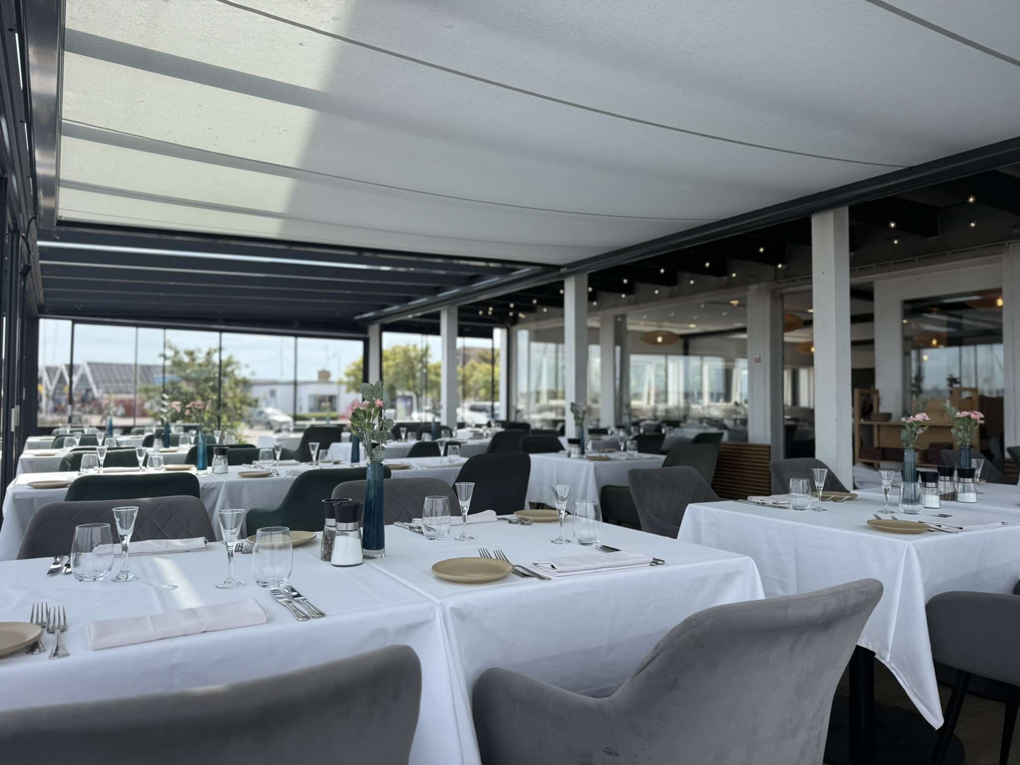 Overdækket terrasse med hvide duge, grå stole og udsigt mod havn