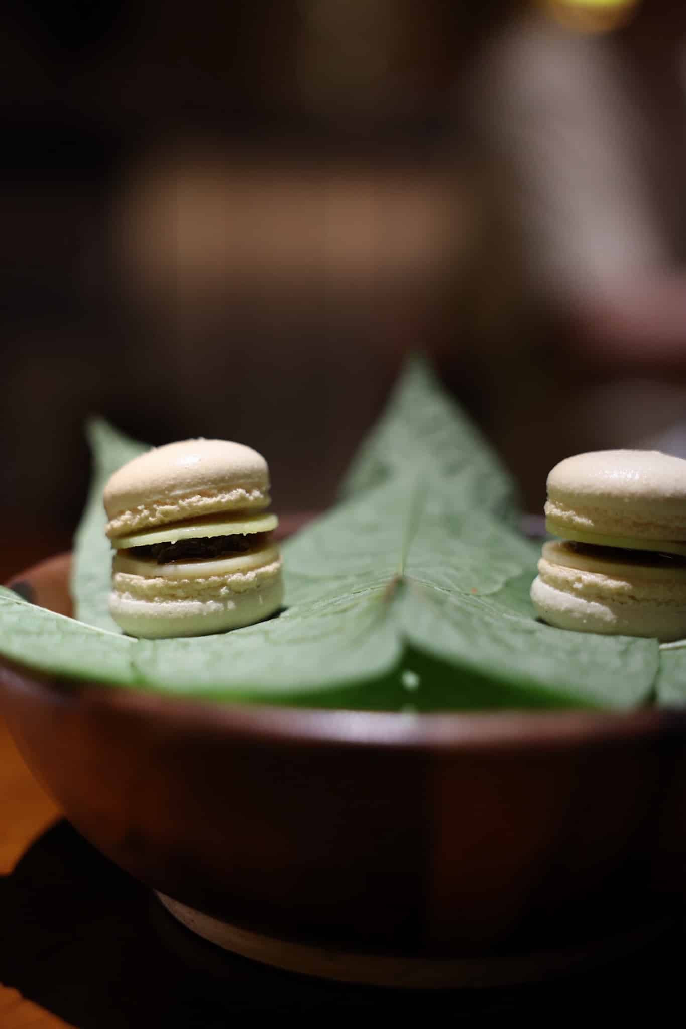 To lyse macarons med mørkt fyld serveret på grønne blade i træskål