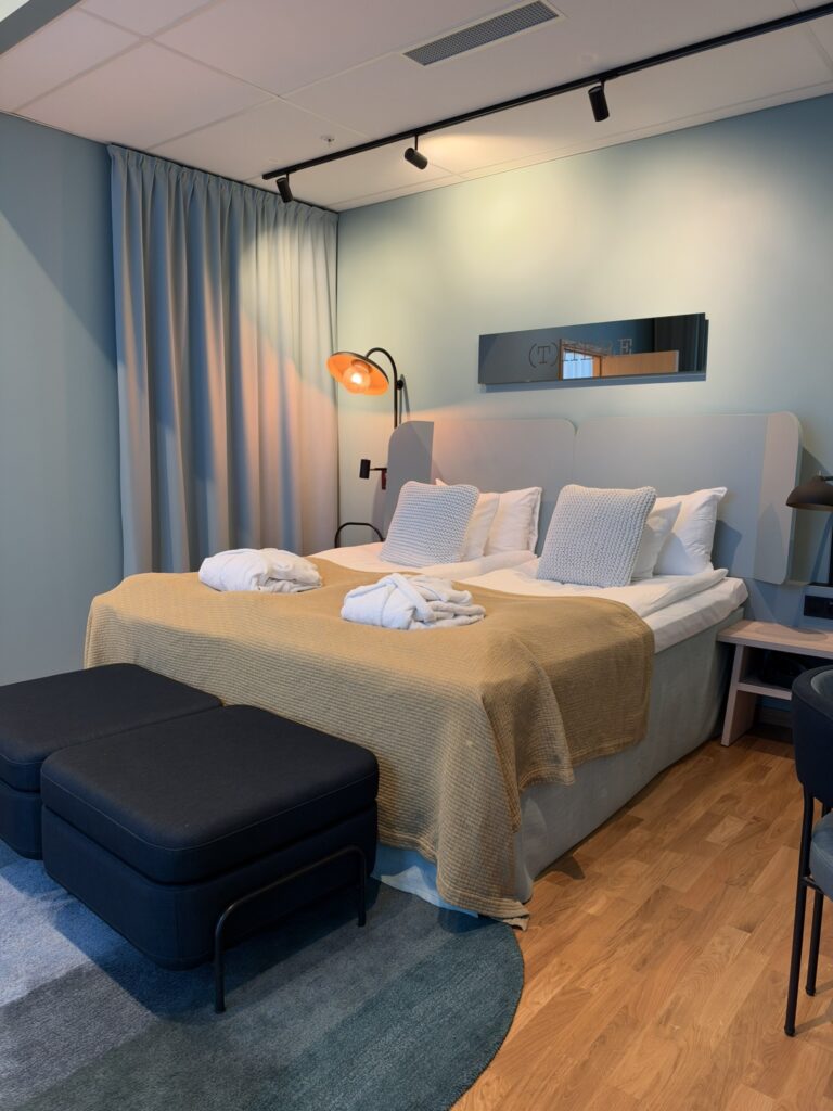 Hotelværelse med dobbeltseng, gult sengetæppe, badekåber og to mørkeblå puffer