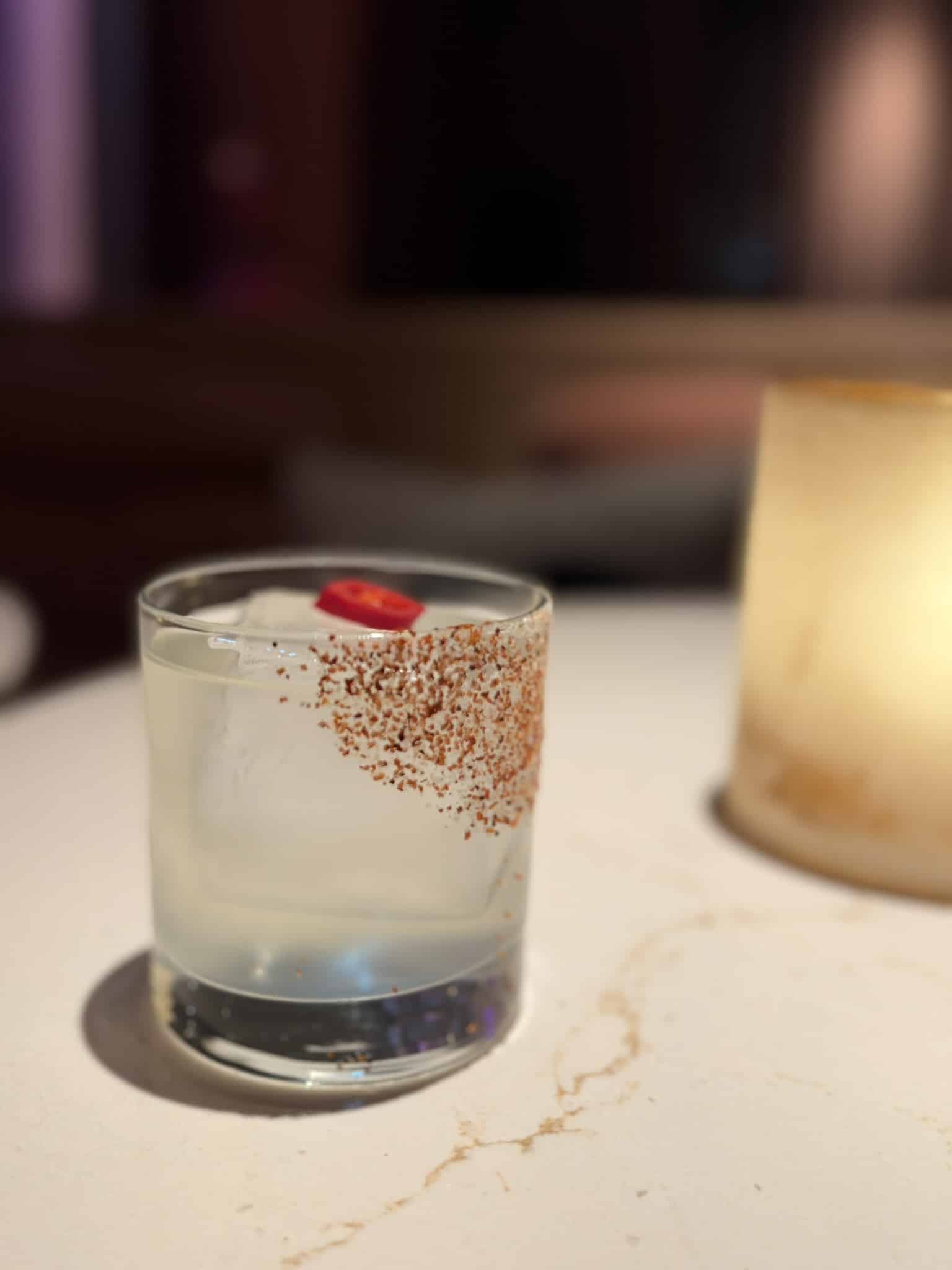 Klar cocktail med stor isterning, chiliskive og krydret saltkant på glasset.