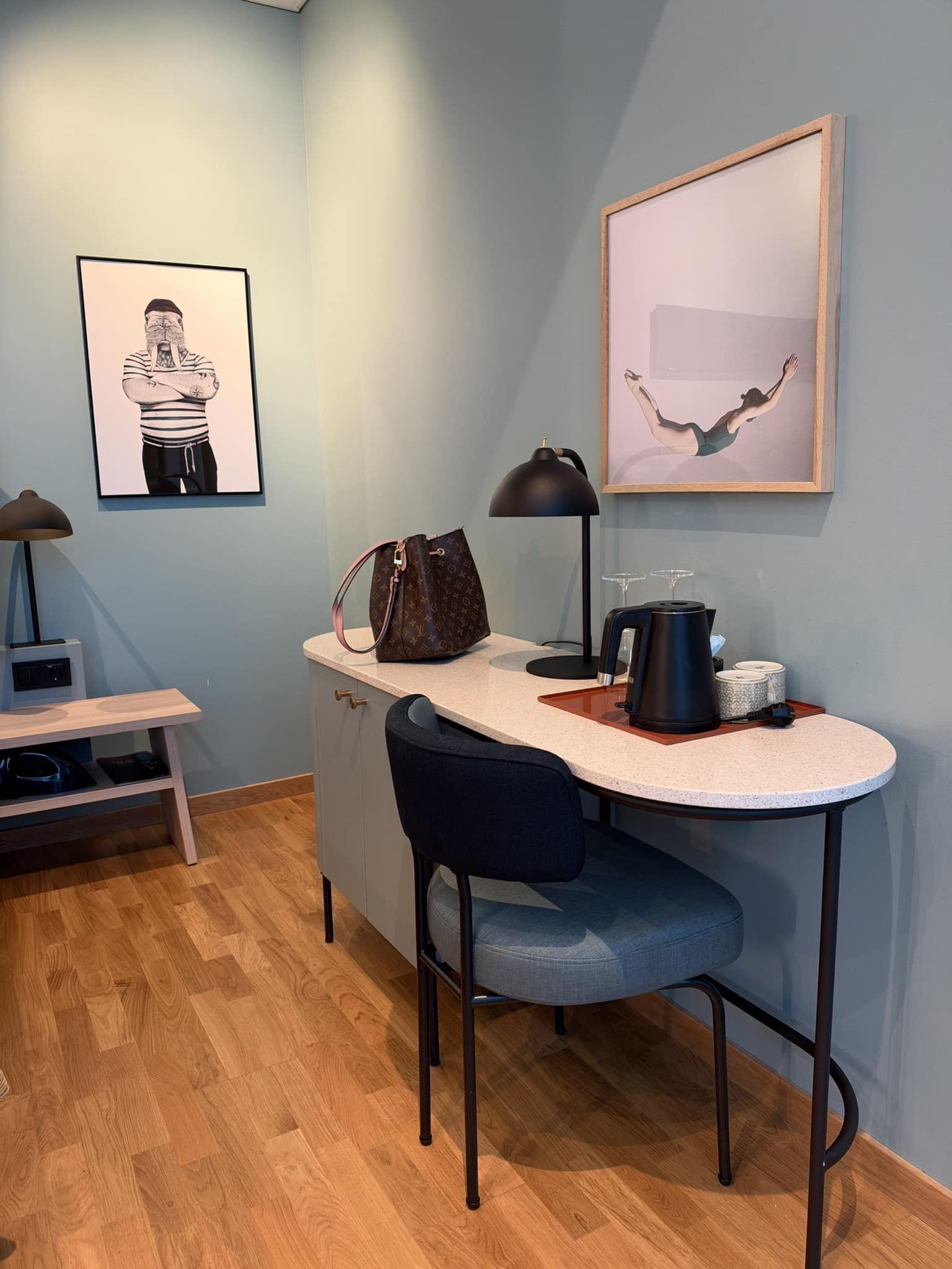 Skrivebord med sort lampe, elkedel, kopper, håndtaske og to indrammede billeder