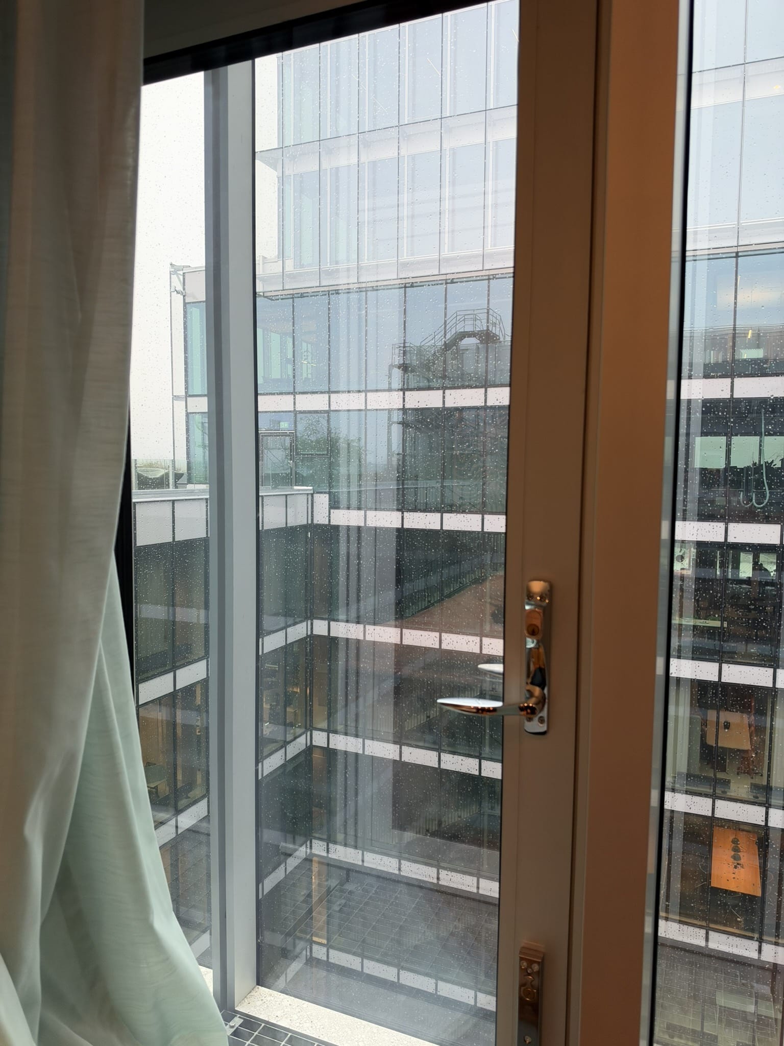Udsigt fra hotelværelsets vindue til glasfacader på nabobygning med regndråber