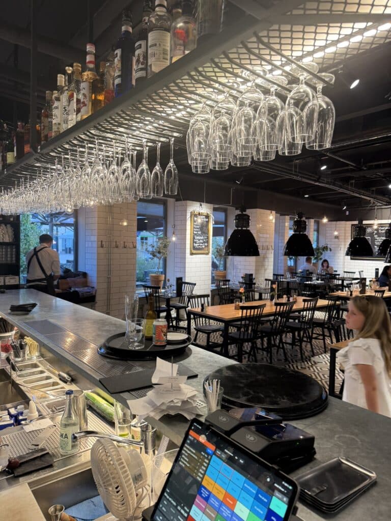 Bar med ophængte glas og spiritusflasker, hvide fliser og sorte stole i restaurant