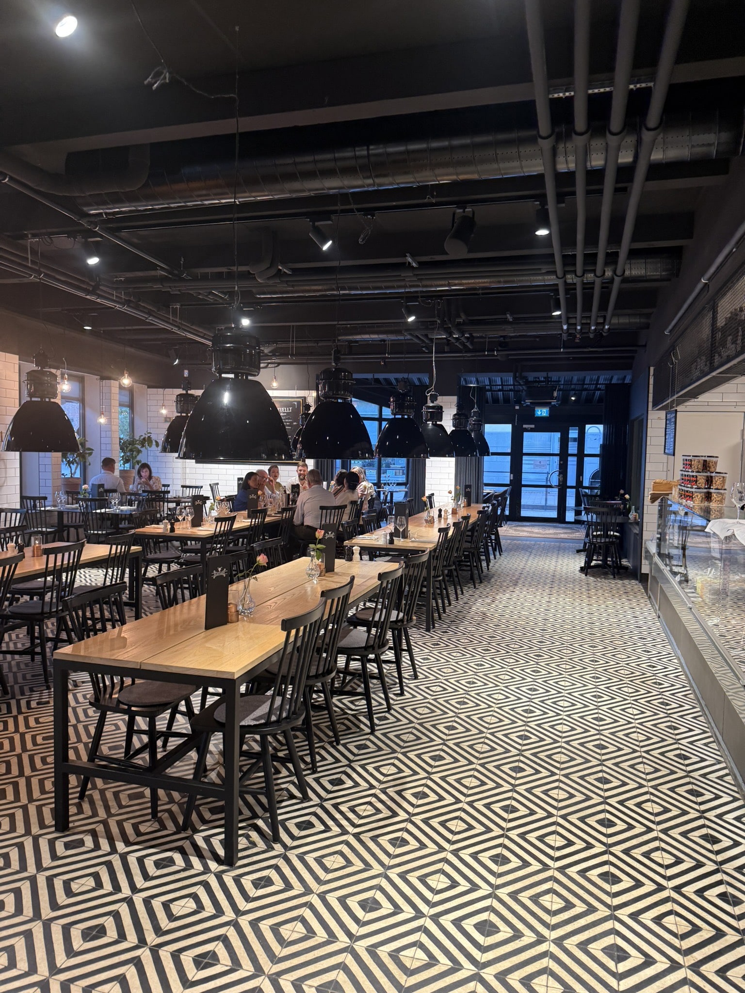 Restaurantlokale med lange træborde, sorte stole, industrilamper og mønstret gulv