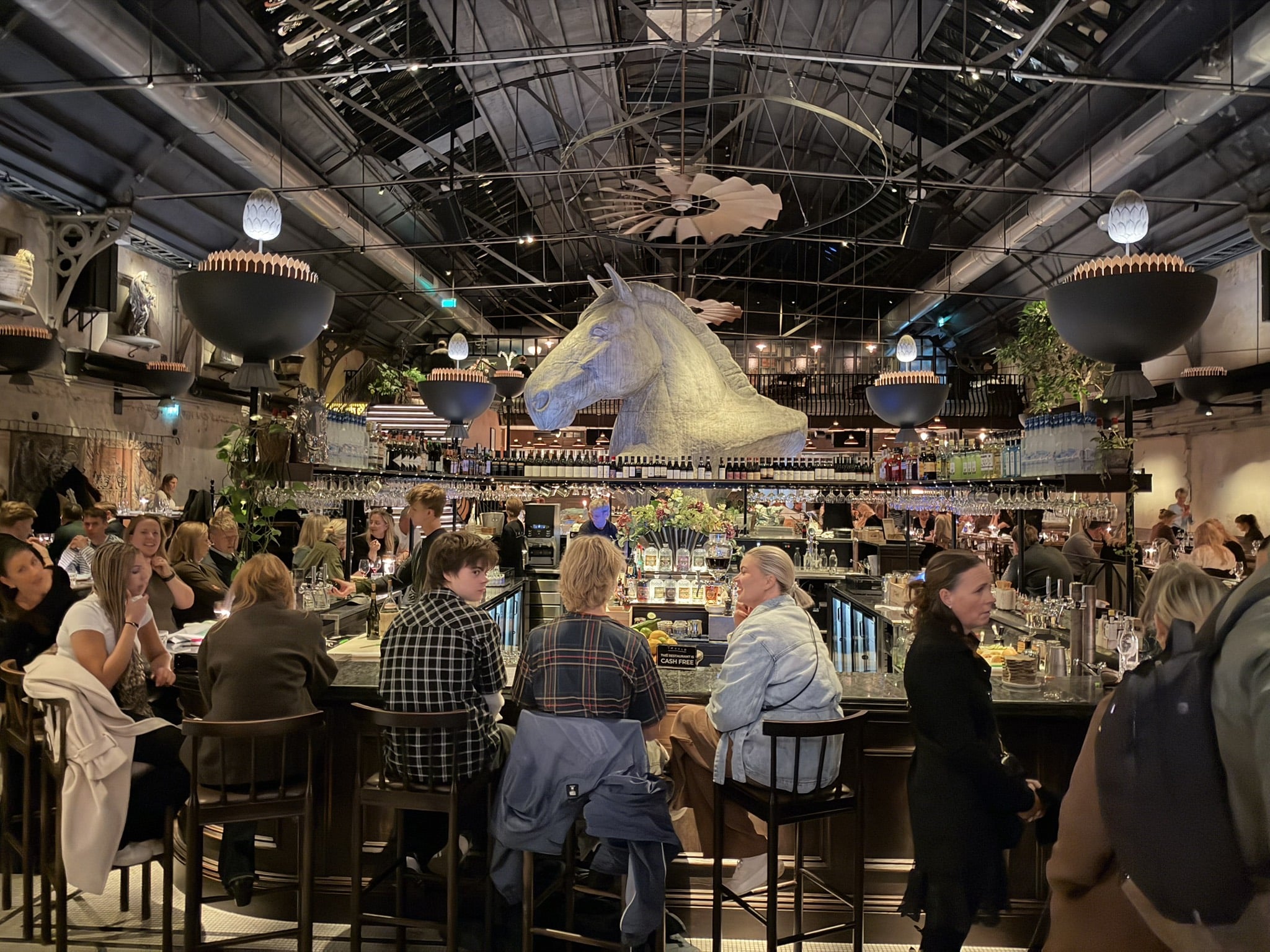 Stor bar med gæster på barstole under et stort hestehoved i loftet.