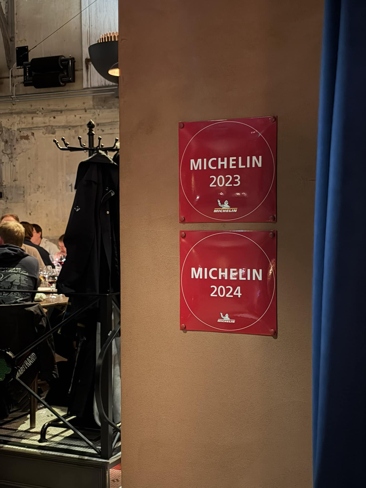 To røde Michelin-skilte for 2023 og 2024 på væg ved blåt gardin.