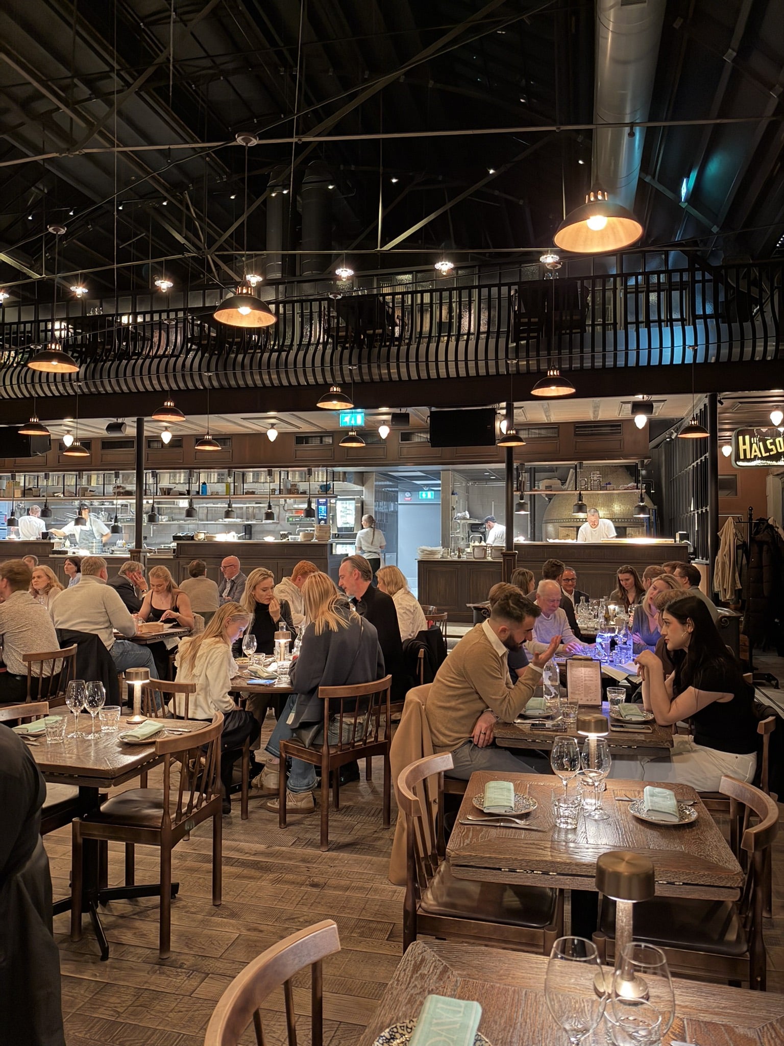 Restaurantsal med træborde, gæster, pendellamper og åbent køkken i baggrunden.