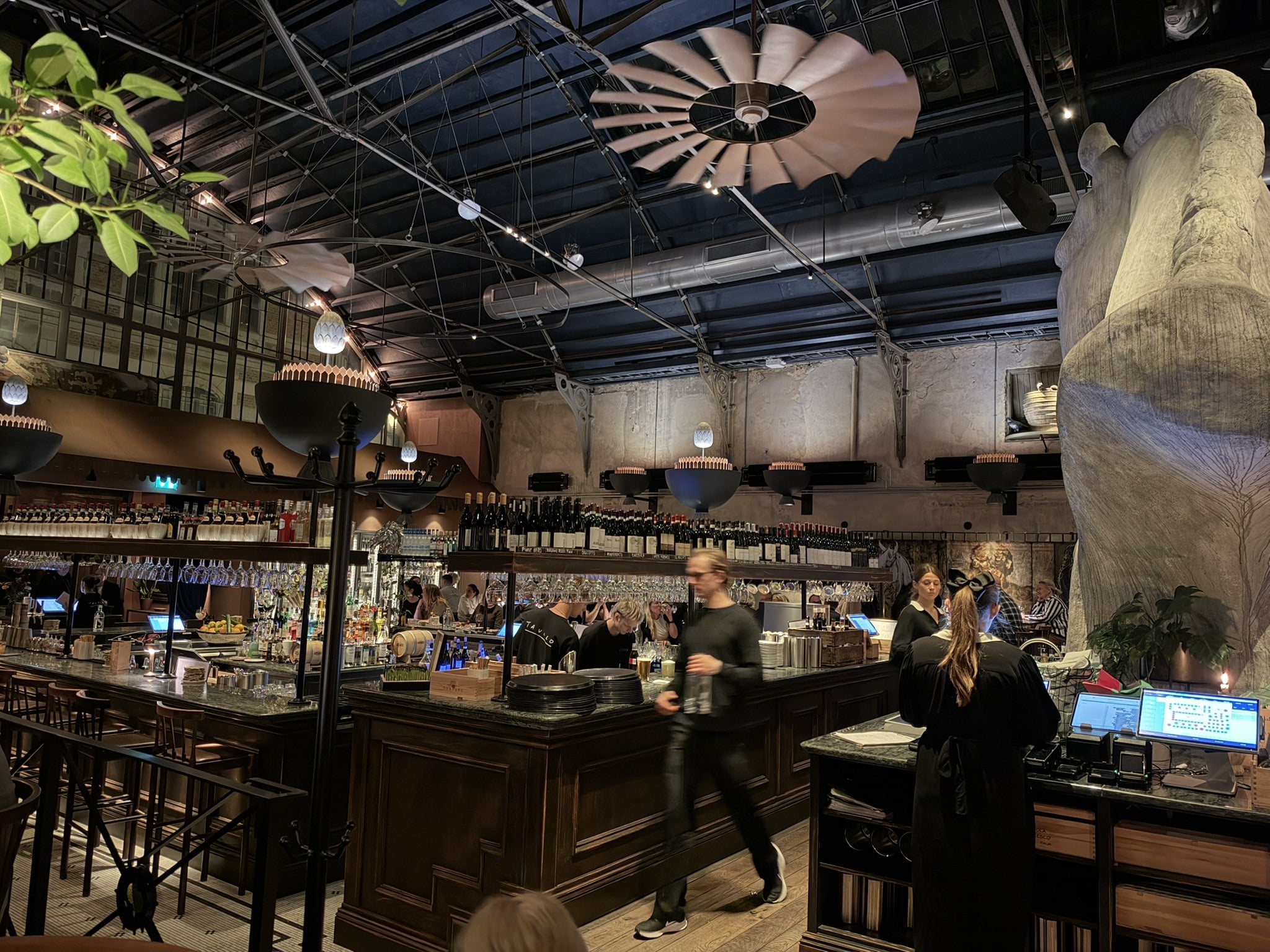 Restaurantlokale med høj bar, vinhylder, loftventilator og stor skulptur